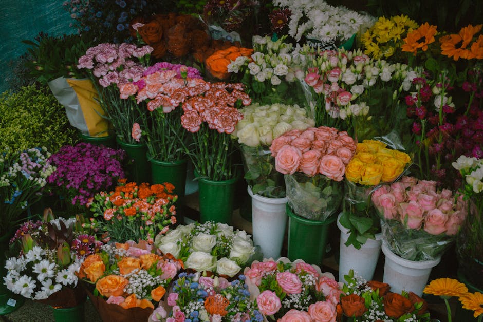 A vibrant display of fresh flowers arranged in buckets and bouquets at a flower shop. The assortment includes pink, white, yellow, orange, and purple blooms such as roses, carnations, daisies, and chr