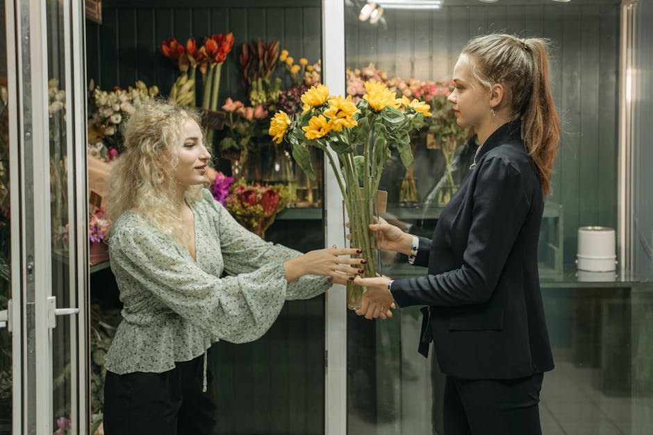 A young woman with curly blonde hair, wearing a light grey blouse with a floral pattern, is receiving a bouquet of bright yellow sunflowers with green stems and large petals from another woman in a da
