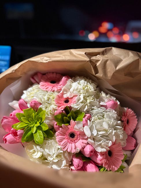 A close-up view of a floral bouquet wrapped in brown craft paper, featuring a mix of white hydrangeas, pink gerbera daisies, and bright green succulents, arranged in a compact, rounded style. The flow
