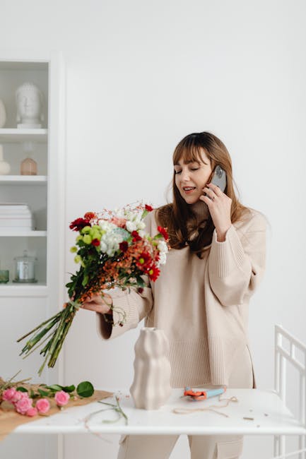 A young woman with long brown hair and bangs is smiling while talking on a mobile phone held to her right ear. She is wearing a beige sweater and holding a vibrant floral bouquet consisting of red, wh
