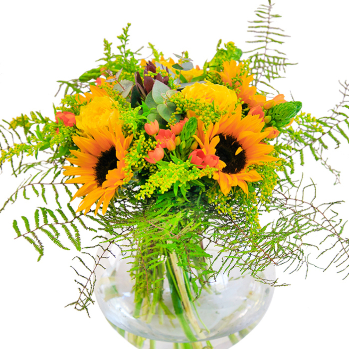 Glowing with warmth and character, this bouquet is a true reflection of Abbey Wood's welcoming community spirit. Towering sunflowers with deep espresso centers dominate the arrangement, their yellow-orange petals arching outward in radiant layers. Flanking the sunflowers, tightly packed yellow garden roses add elegance and their own gentle lustre, reminiscent of the gardens near Lesnes Abbey. Rich green ferns and light, cascading vines spill gracefully among the blooms, giving the design a sense of movement-like a breeze sweeping through Bostall Woods. Punctuating the verdant display, bright red Hypericum berries shine with playful energy, their smooth surfaces contrasting beautifully with the textural petals around them. Pale goldenrod sprigs and slender green spikes contribute hints of meadow charm. Subtle eucalyptus leaves in muted blue-green enhance the arrangement's rustic, natural feel. All are expertly presented in a clear, rounded glass vase, exposing the bouquet's vibrant stems in crystal-clear water. The bouquet carries a gentle, uplifting scent, evoking summer walks and garden celebrations across Abbey Wood. Perfect for birthdays, anniversaries, or as a joyful centrepiece, this bouquet from Flower Delivery Abbey Wood is crafted to bring happiness and optimism for every cherished occasion.